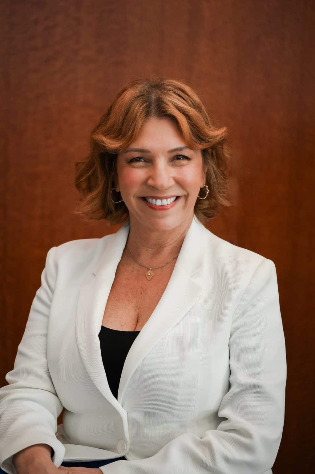 A professional headshot of a smiling woman with wavy auburn hair wearing a white blazer and a gold necklace against a warm brown background.