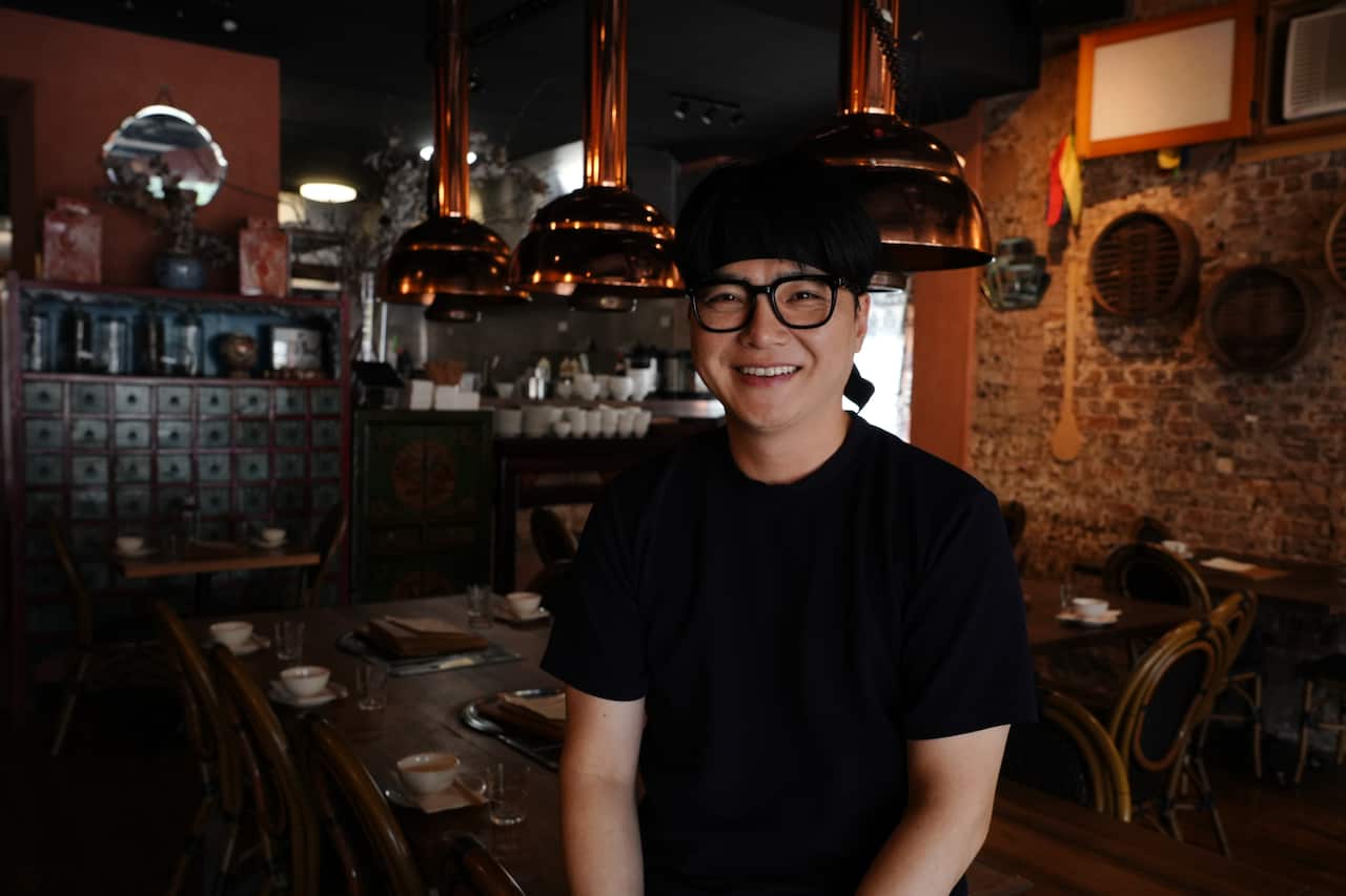 A Korean man wearing a black t-shirt smiling warmly and looking at the camera. In the background are copper coloured smoke vents hanging from the ceiling with dark wooden restaurant tables and chairs, set with bowls and plates.
