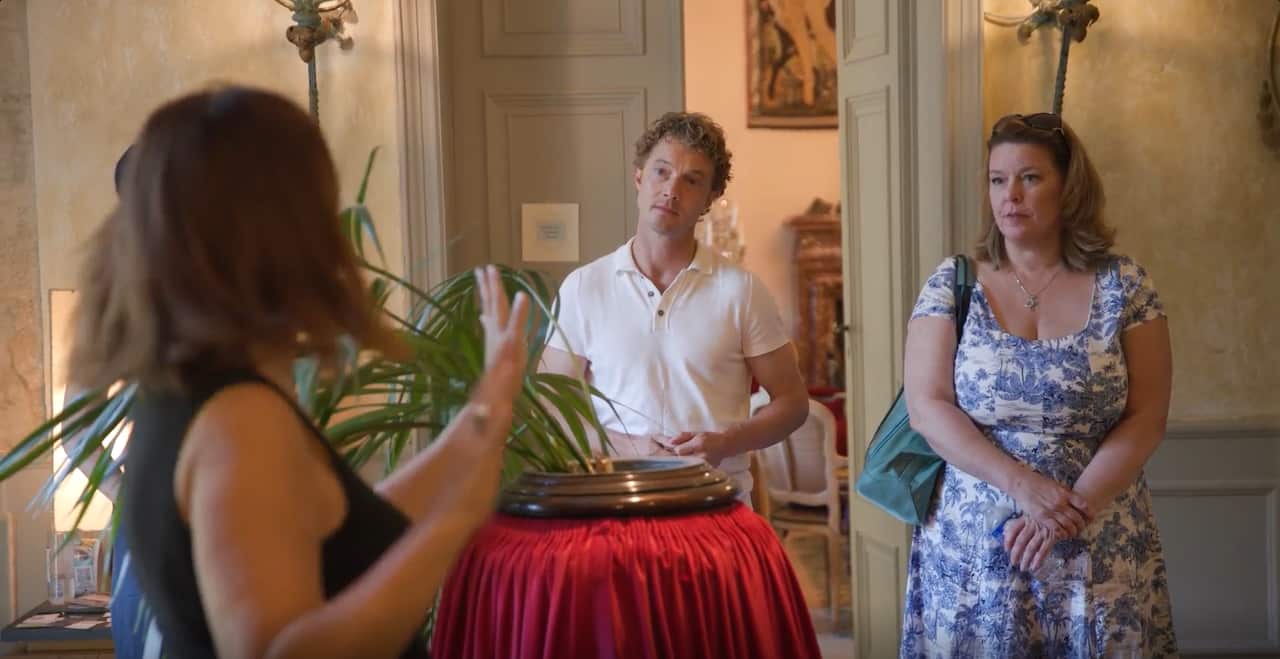 A man and a woman listen to another woman speaking. They are standing inside a room of a chateau.