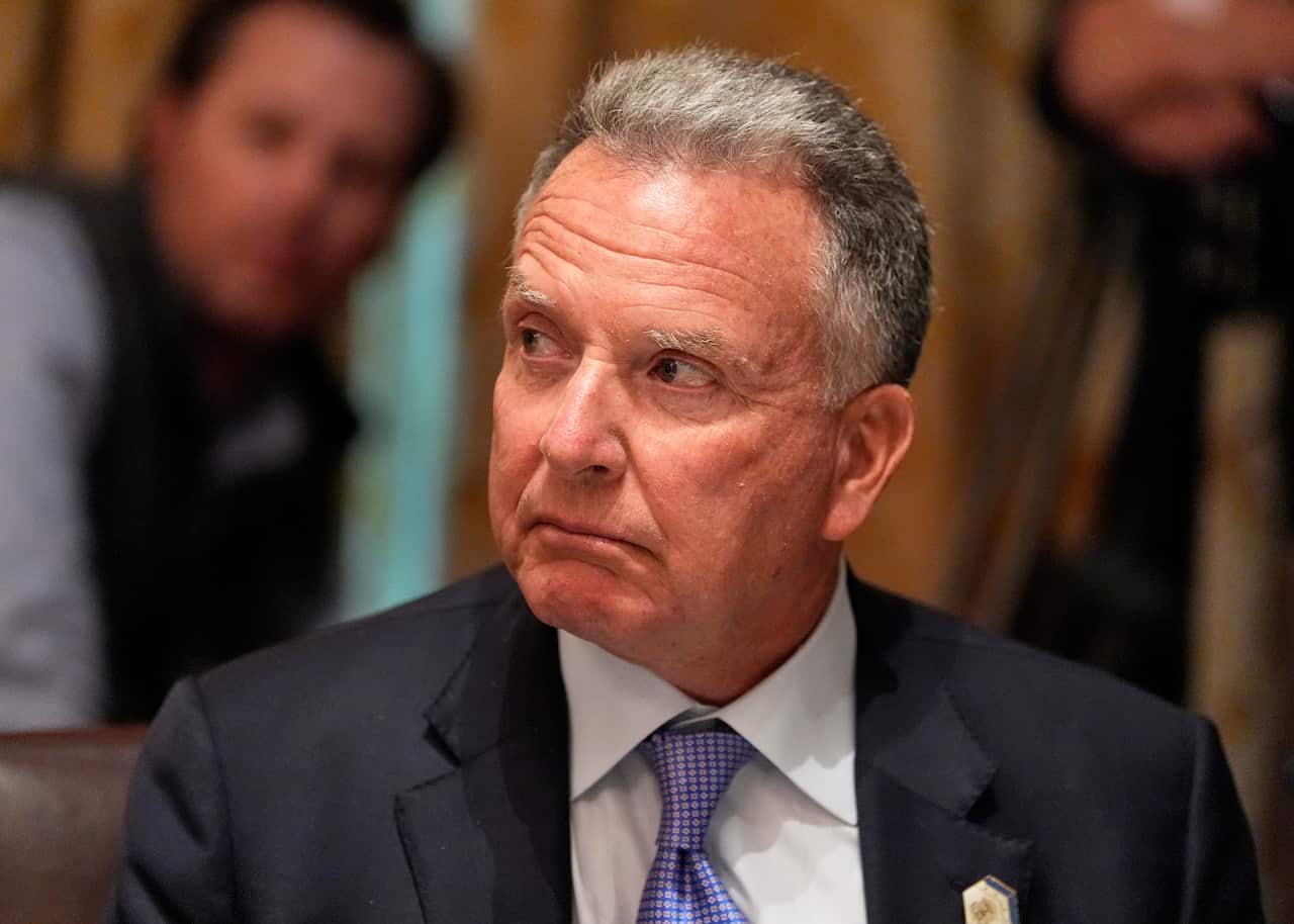 A man with grey hair, wearing a black blazer, white shirt and purple tie.
