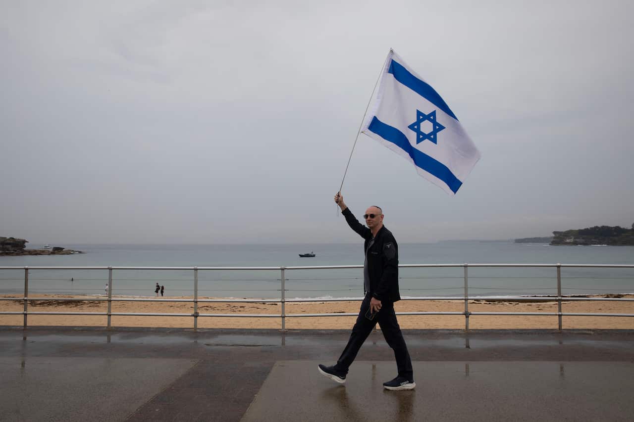 A man holding an Israeli flag as he walks along a promenade, a beach is in the background. 