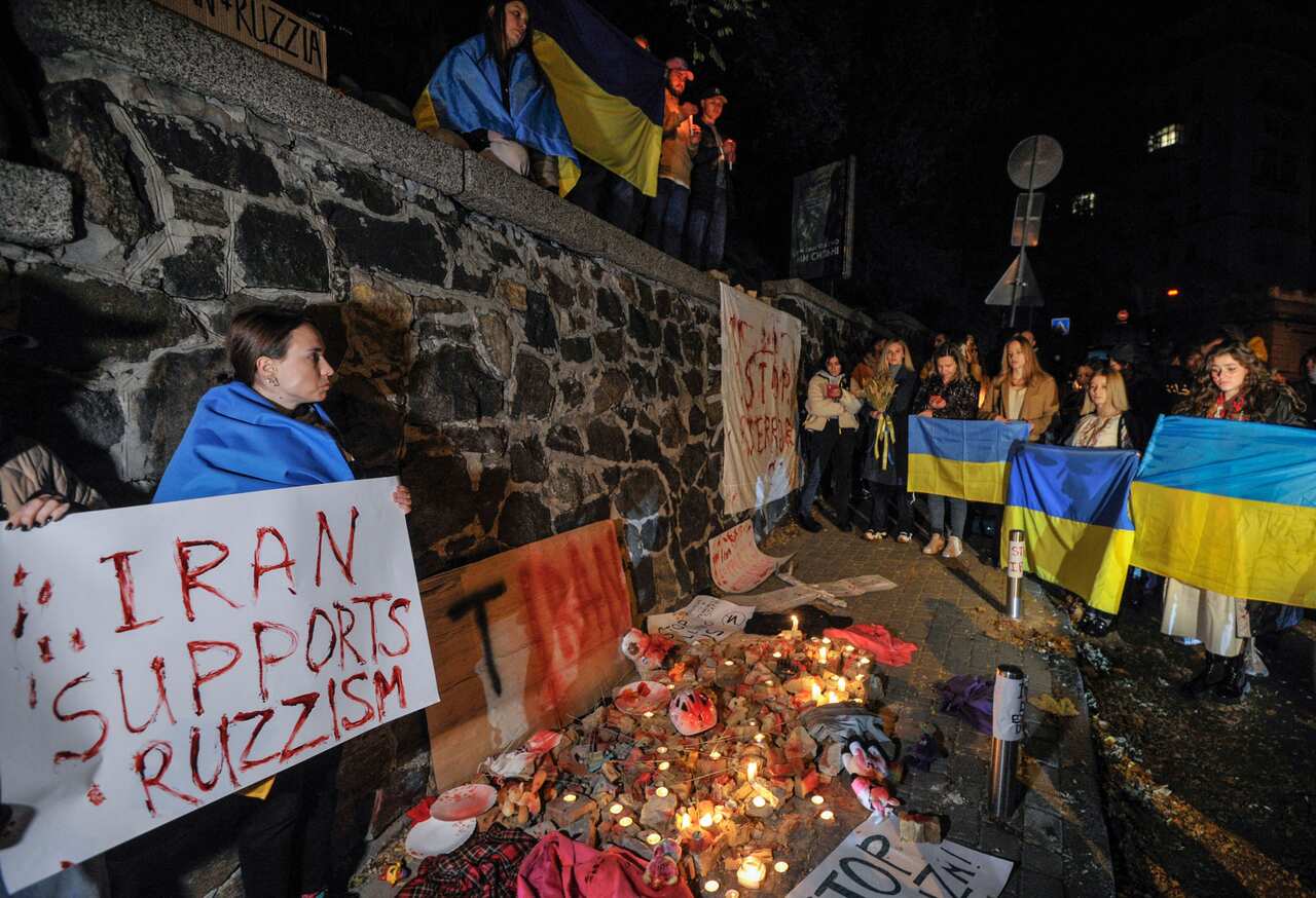 Two people hold up a sign reading "Iran supports Ruzzism". Near them are a group of candles and several people holding up Ukrainian flags. 