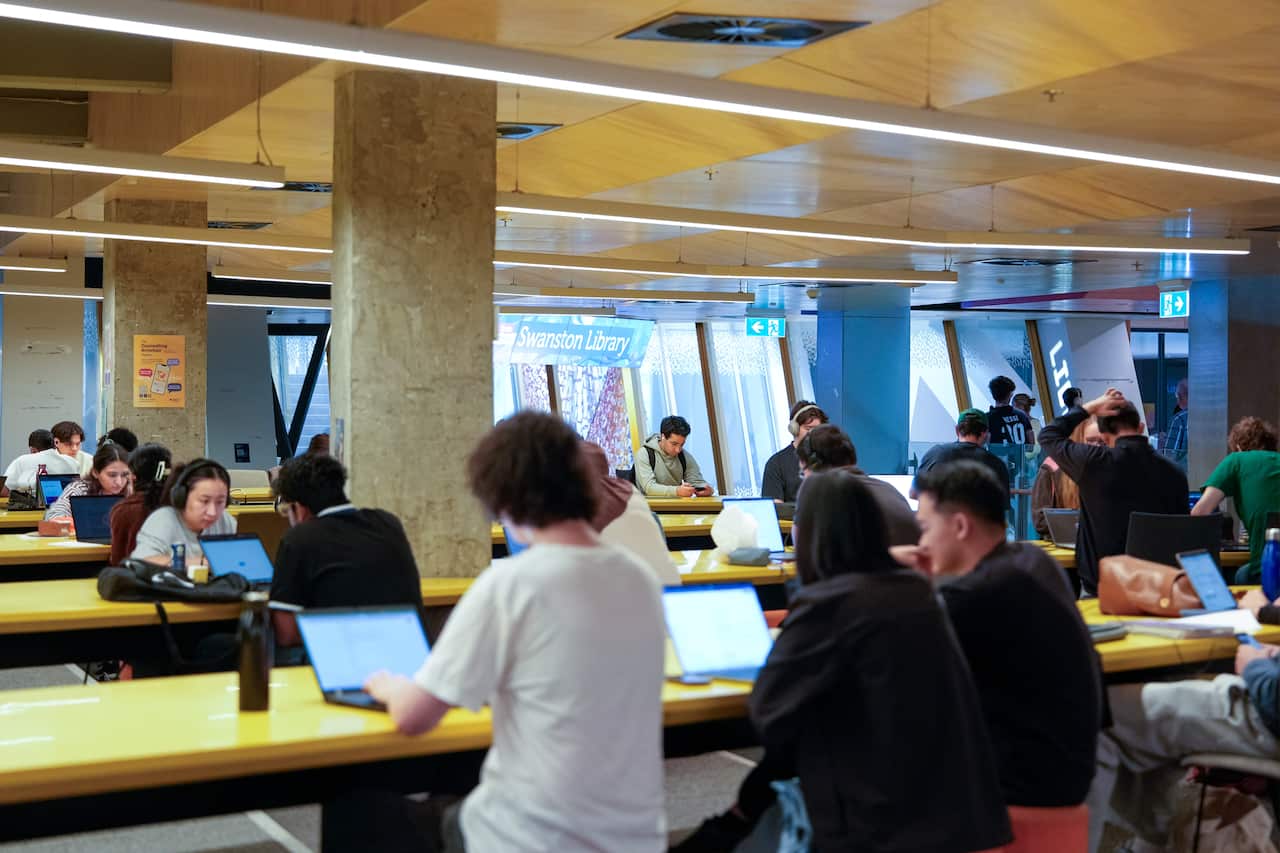 University students sitting in front of laptops on long wooden desks in a hall.