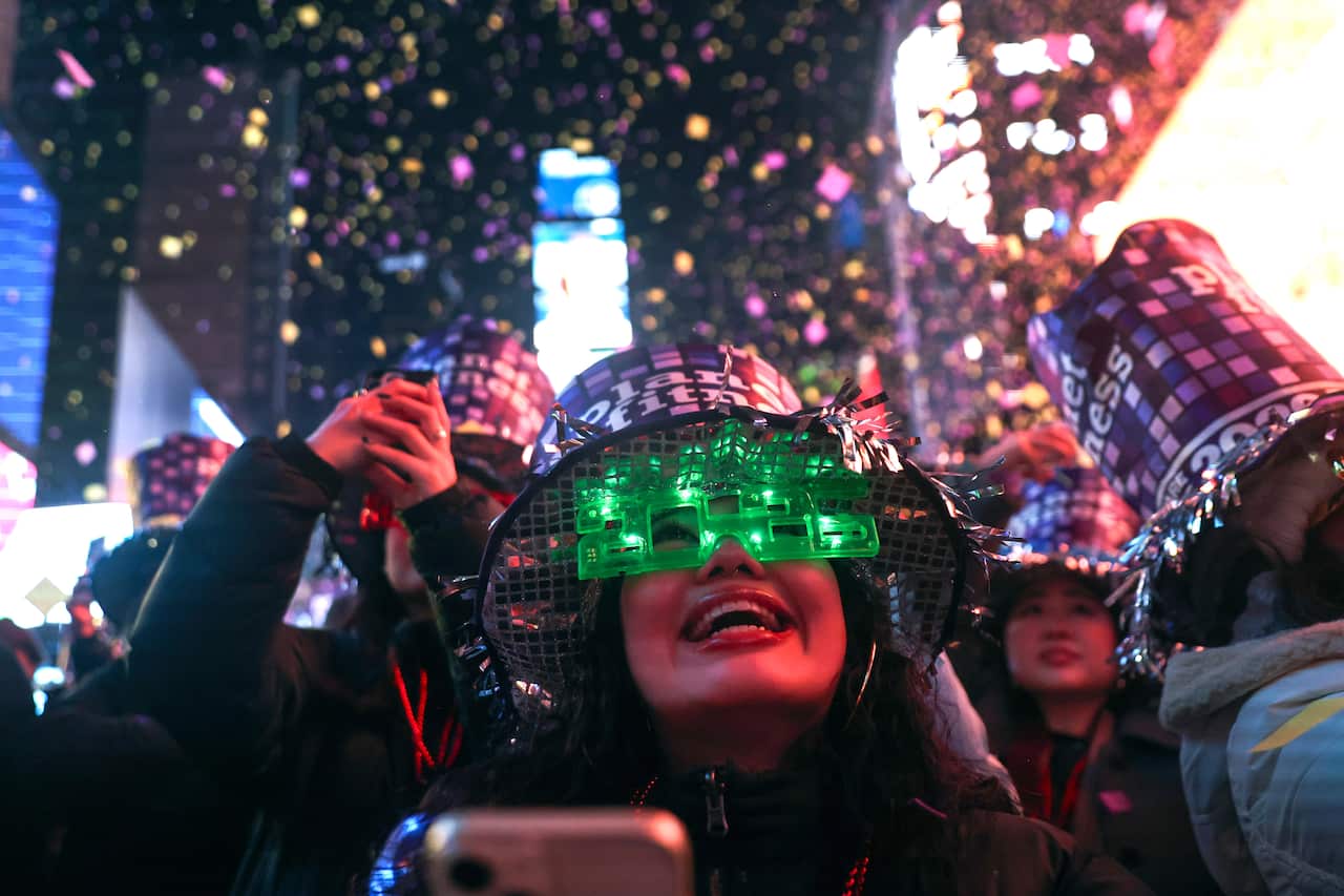 A joyful reveller wearing glowing green "2026" glasses and a Planet Fitness hat smiles as colourful confetti falls during a New Year's Eve celebration in Times Square.