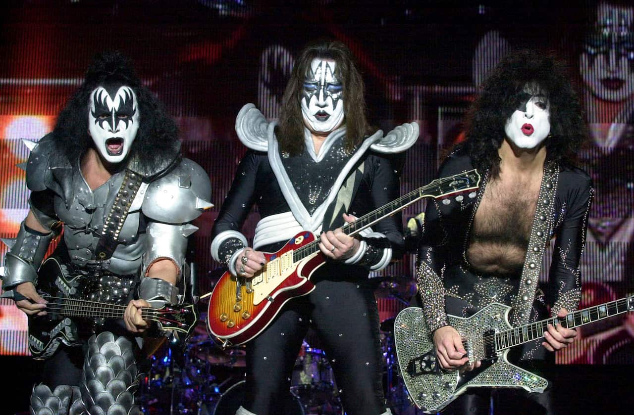 Three guitarists with face paint and elaborate costumes perform on a stage.