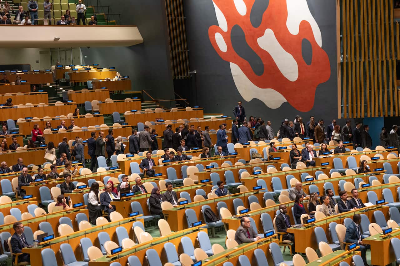 A wide, slightly elevated photo shows delegates walking out of the United Nations General Assembly Hall, while in largely empty rows a few attendees are seated at wooden desks with blue chairs. 