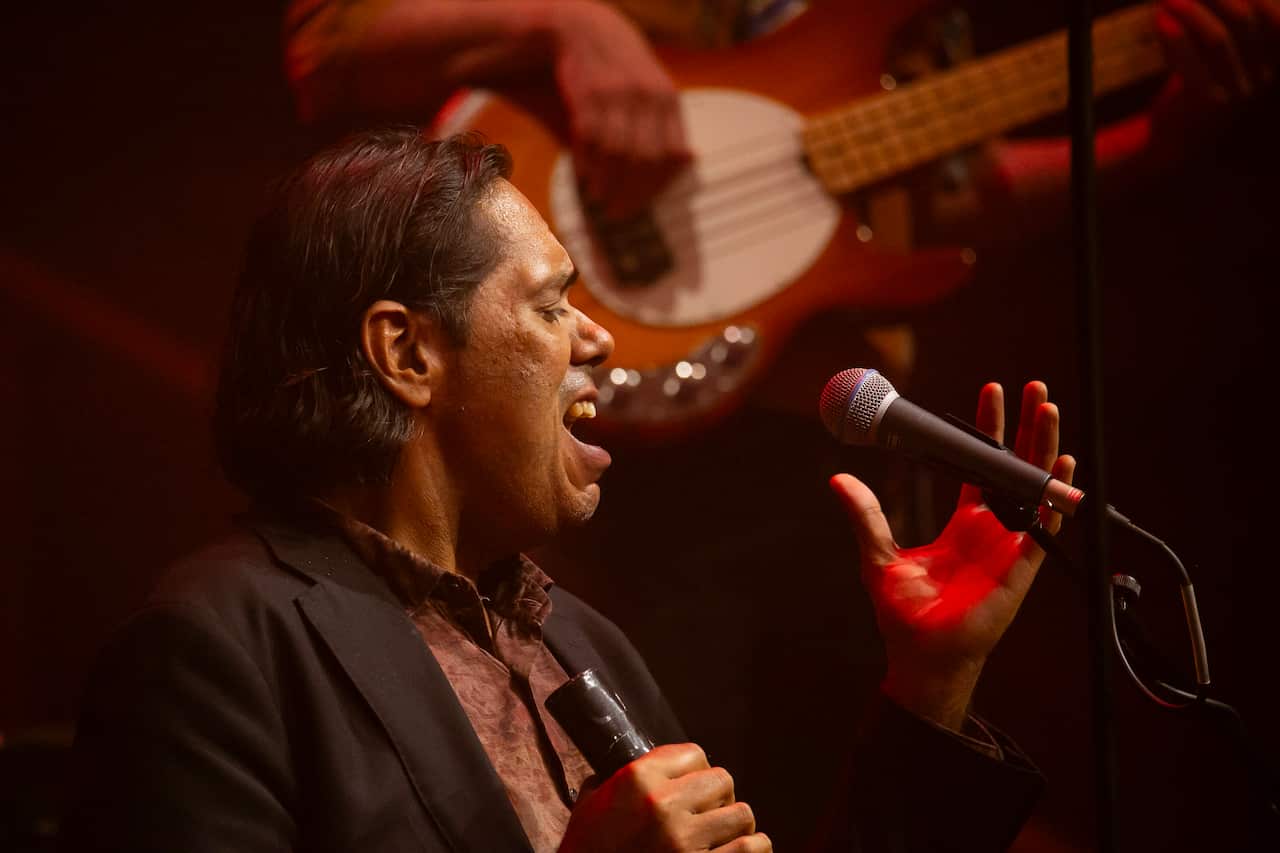 A man passionately sings into a microphone on stage, with a bass guitar player in the background.