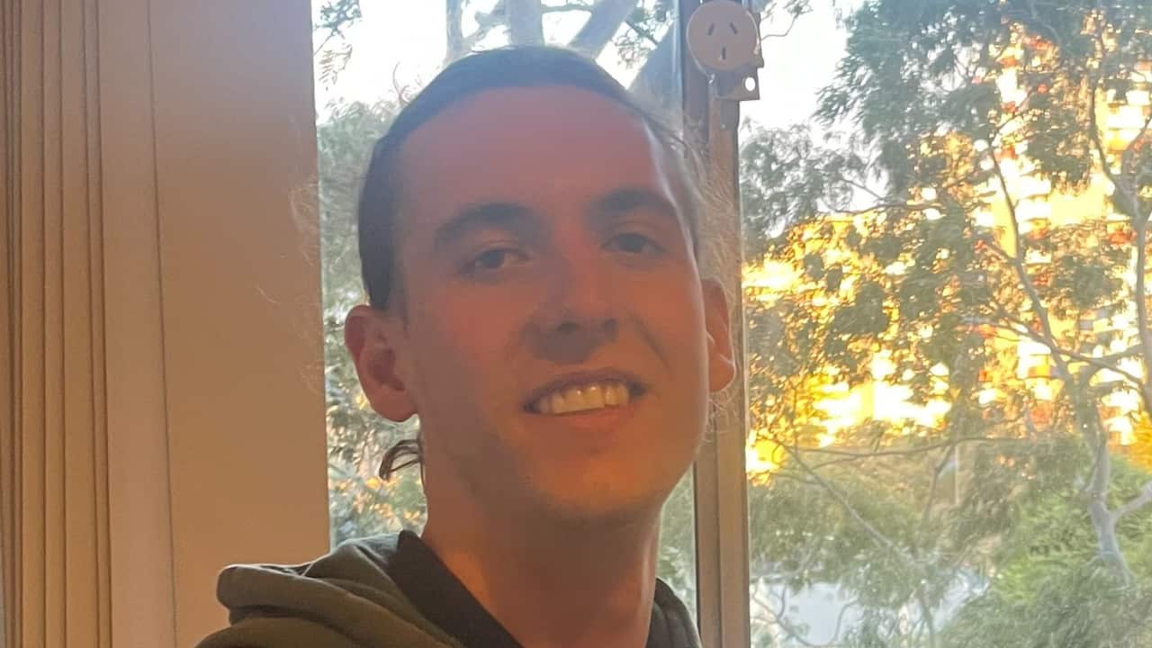 a close up of a young man with a ponytail and wearing a hoodie stands in front of a window with sunset light and trees in background