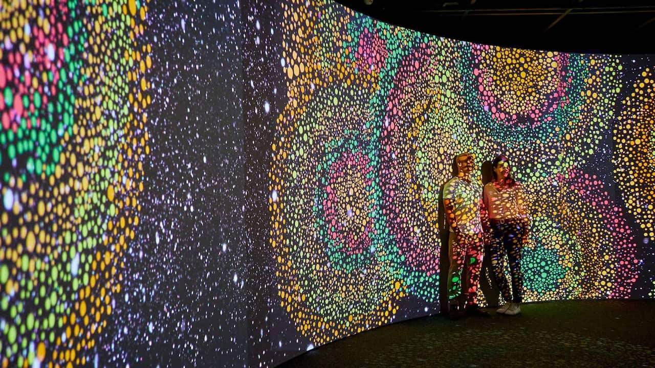 Two people stand in front of Aboriginal art projected on a wall