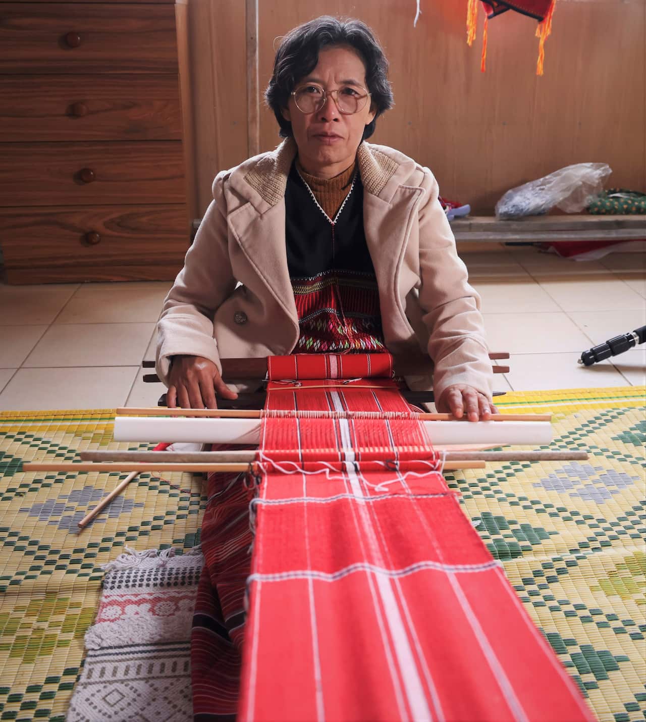 A woman in a beige jacket sits on the floor, weaving a length of red fabric on a hand-held loom.