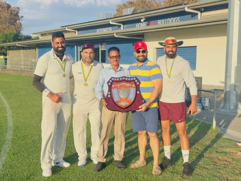 Five men stand together on a cricket oval in front of a small building, with three of the men wearing medals around their necks and two men holding a large ceremonial shield.