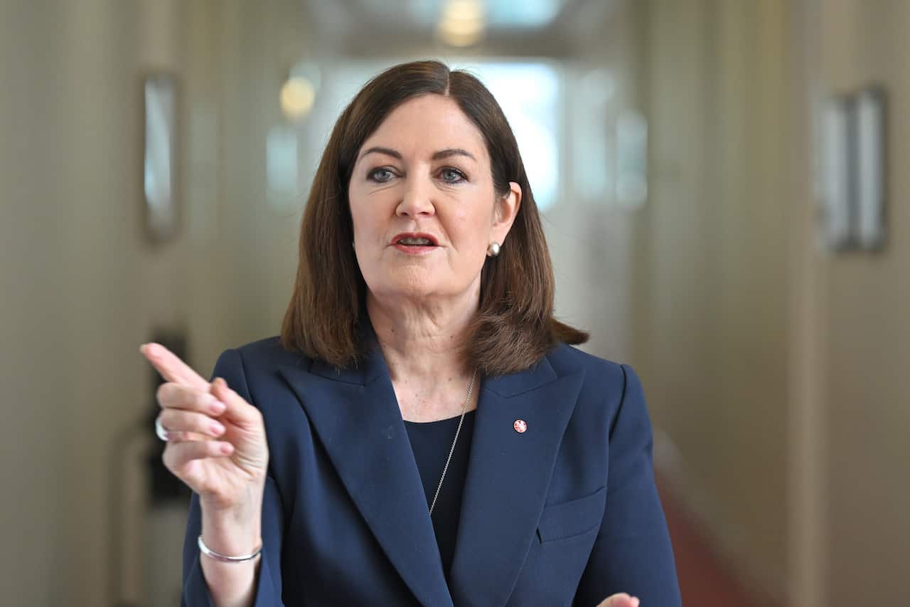 A woman in a suit is pointing her finger while speaking in a corridor.