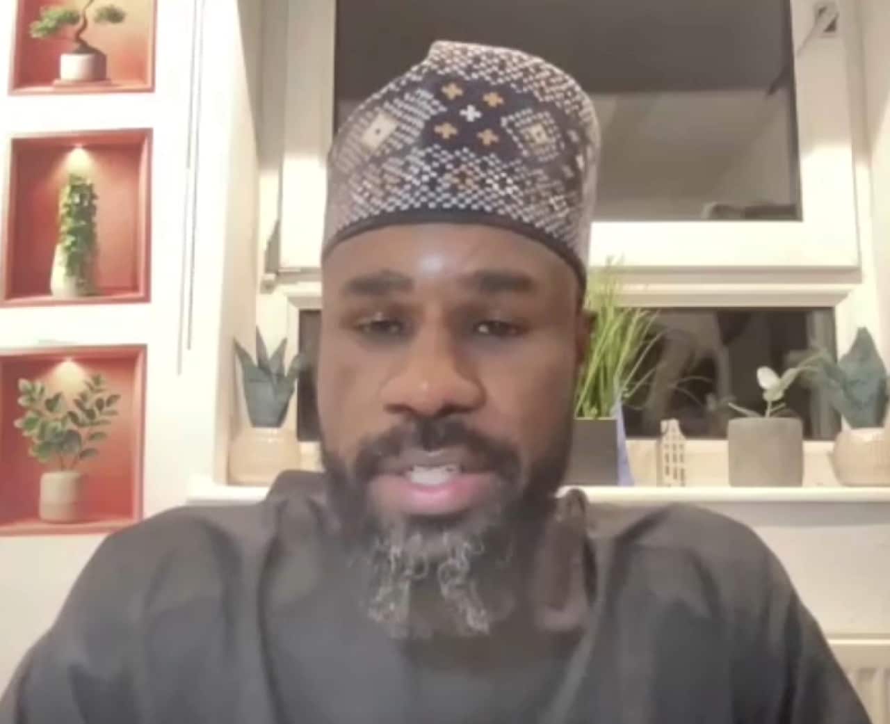A man with a dark beard and a patterned hat sits in front of a white wall featuring small plants in recessed shelves.