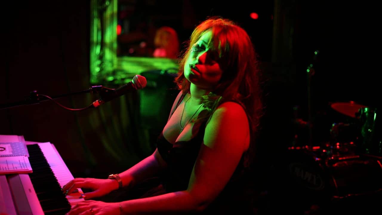 a young woman with long hair singing at a piano in a dark, neon-lit club