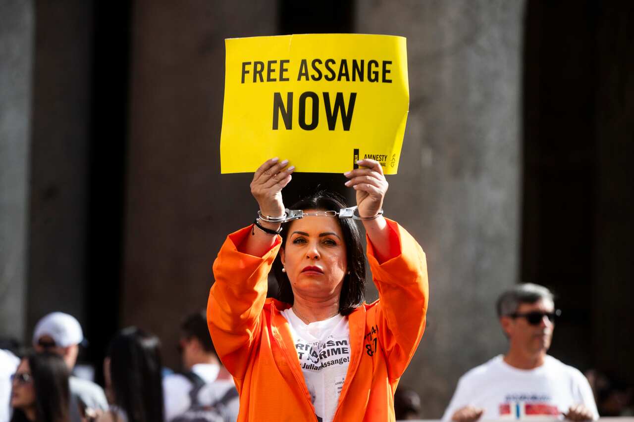 A person in an orange jumpsuit holds up a placard that says 'free Assange now'. They are wearing handcuffs