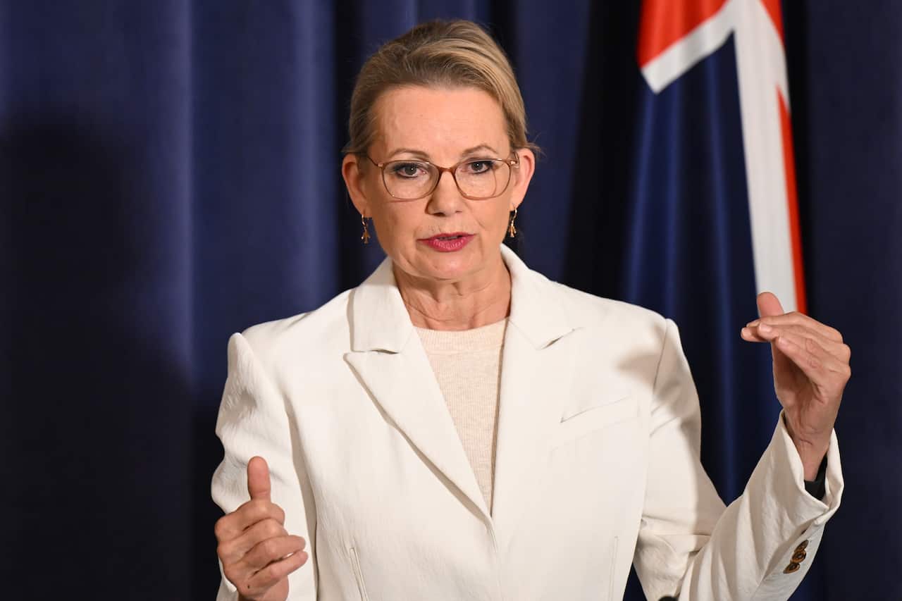 A woman in a white suit addresses the media in front of an Australian flag.
