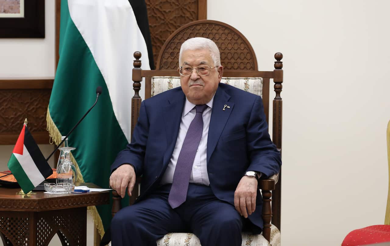 A man in a blue suit sits in a throne-like chair next to a flag.