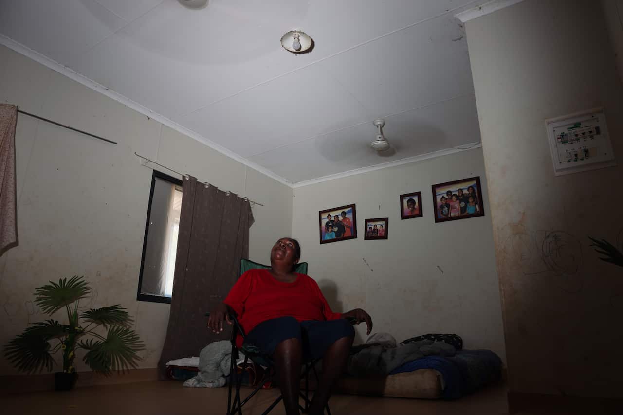 A woman with a red top sits on a camping chair inside a dark room, with the ceiling fan going.