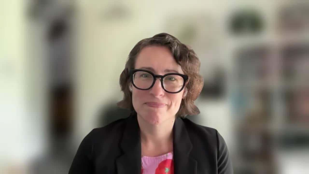 A woman with short brown hair and black-framed glasses smiles while wearing a black blazer over a pink floral top against a blurred background.