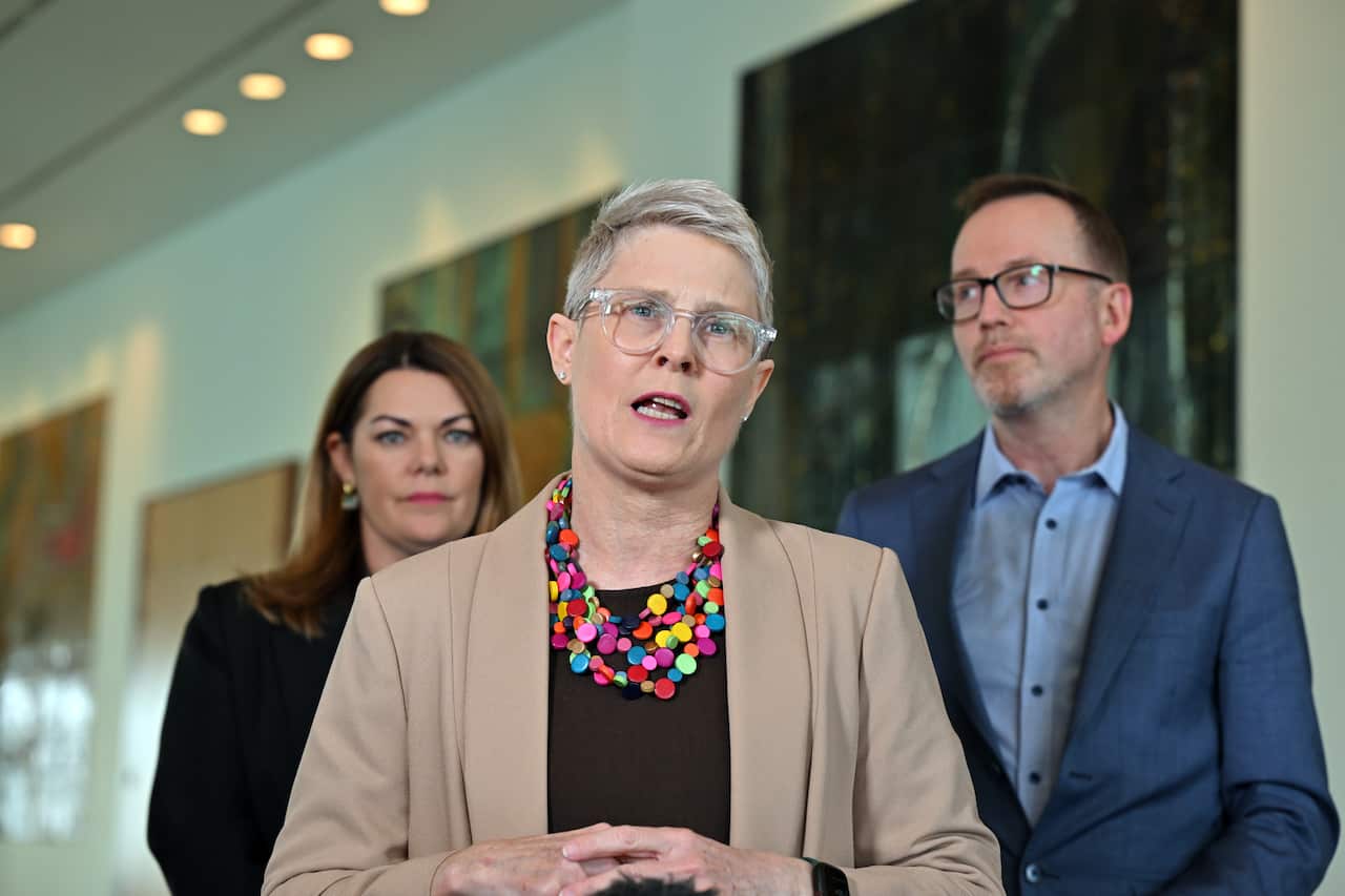 Uma mulher de cabelos curtos, vestindo uma jaqueta bege e um colar colorido na frente de duas pessoas ao fundo.