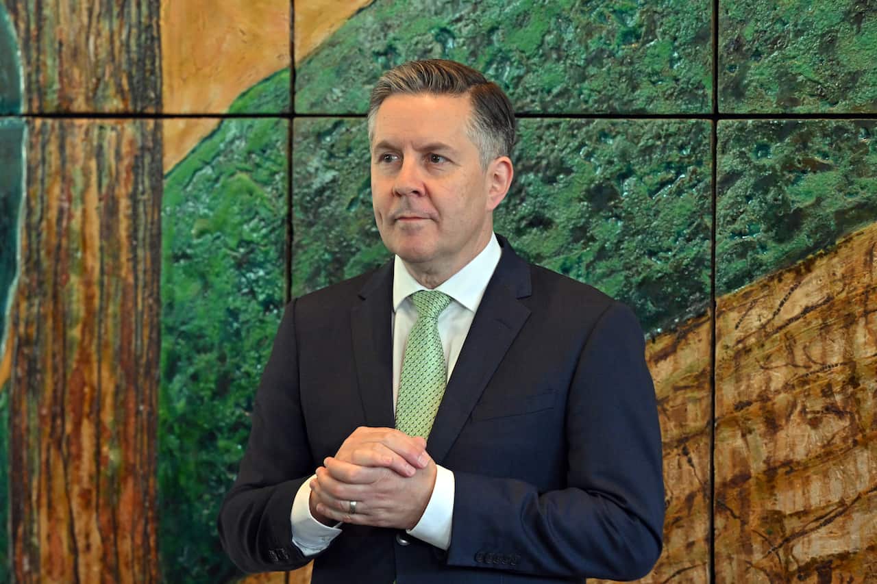 A man in a black suit, white shirt and green tie, standing with his hands folded, in front of a colourful mural.