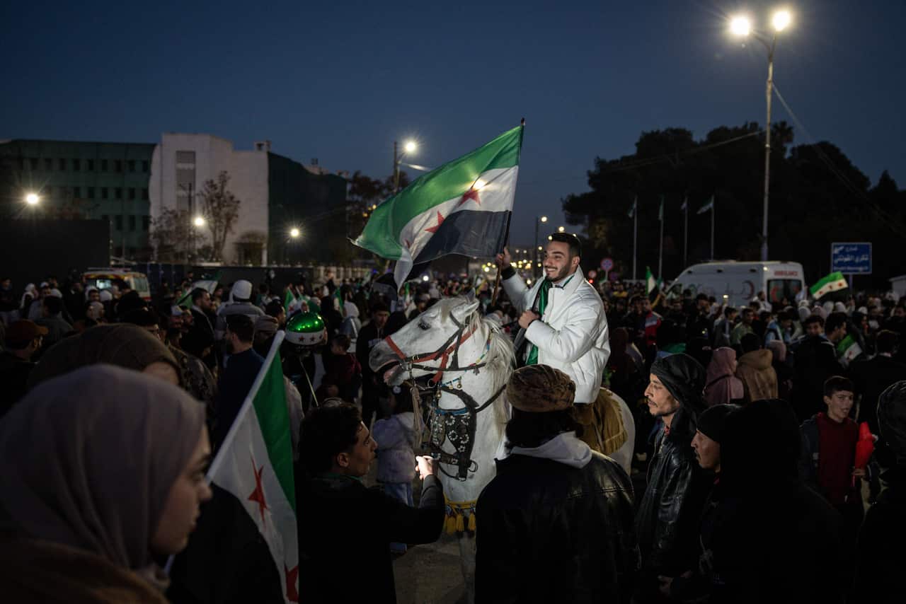 A man on a white horse waves the green, white, and black Syrian revolutionary flag amidst a large, nighttime crowd celebrating in the streets.