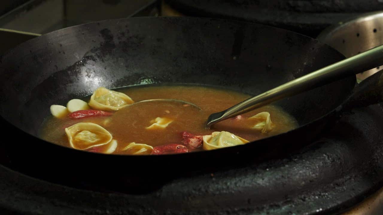 A close up of rice cake soup cooking in a wok on the stove. Meat, dumplings and rice cakes can be seen in the brown broth.