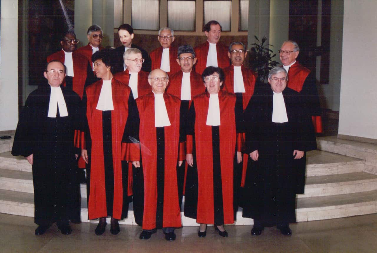 A group of people posing for a photo. Most are wearing judges' robes of black with a red sash on either side of the body and a white legal bib. The two men in front are wearing all black with a white legal bib.