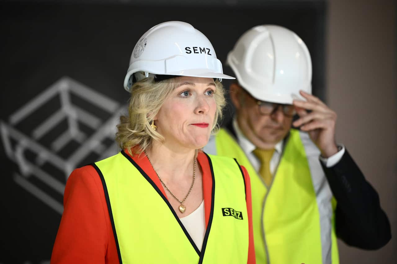A woman in a yellow vest and white construction hat speaks