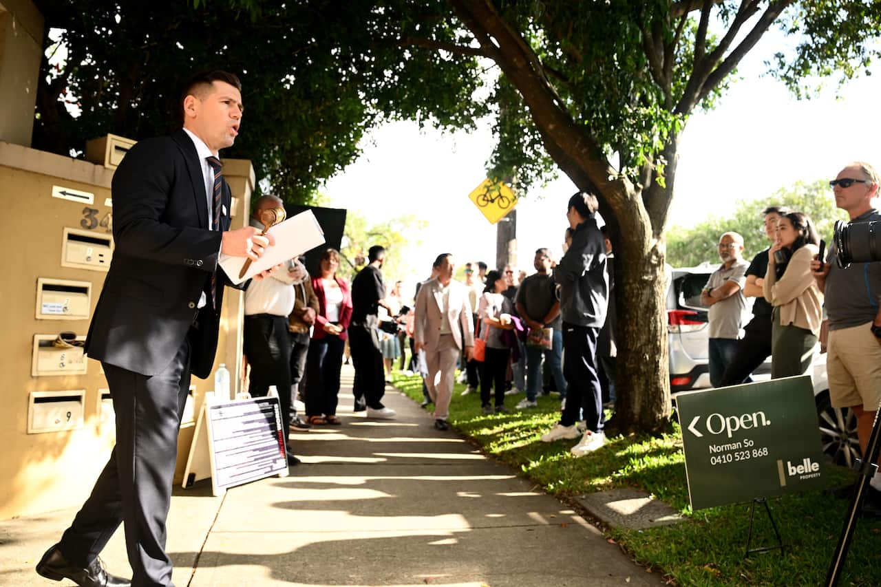 A man in a suit on a footpath shouting at a crowd of other people standing on grass.
