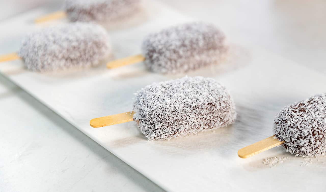 Two rows of chocolate and coconut coated icecreams, with wooden paddle pop stickes, sit on a white platter.
