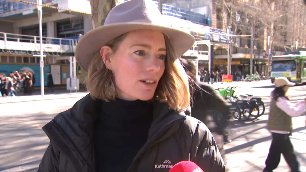 A woman wearing a black jacket speaks into a microphone as a tram passes behind her.