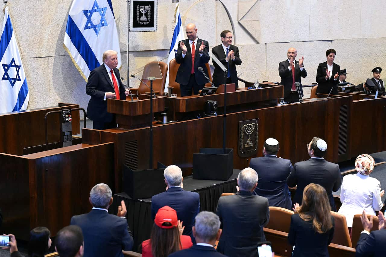 Donald Trump speaking in a parliament room with Israeli flags on the wall. Many of the politicians inside the room are standing and applauding.