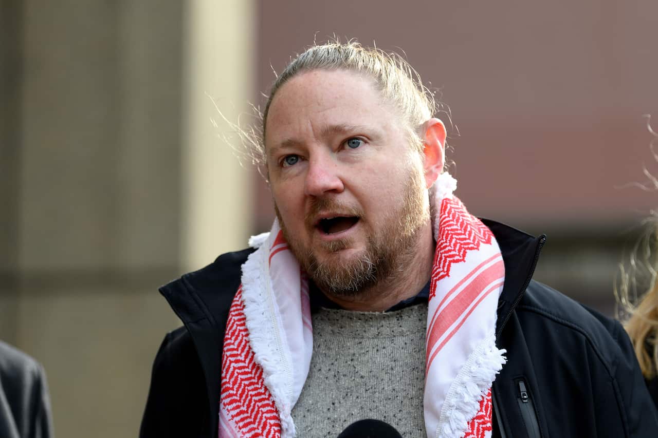 A man speaking outside. He is wearing a keffiyeh scarf around his neck. 