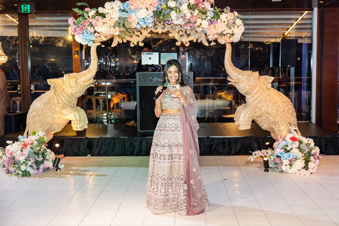 A smiling woman in a heavily embroidered Indian traditional outfit stands on a white dance floor, holding a microphone and a phone, beneath a floral arch supported by two large carved elephant statues at an indoor wedding reception.