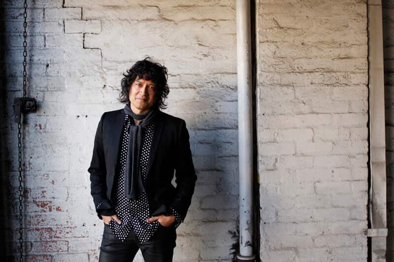 a man with dark curly hair in a black blazer and grey scarf with his hands in pocket stands in front of a white brick wall