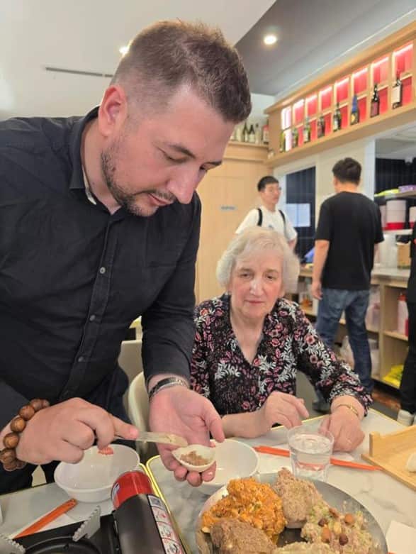 A man in a black shirt stands at a table spooning dumpling filling into a skin while a woman looks on.