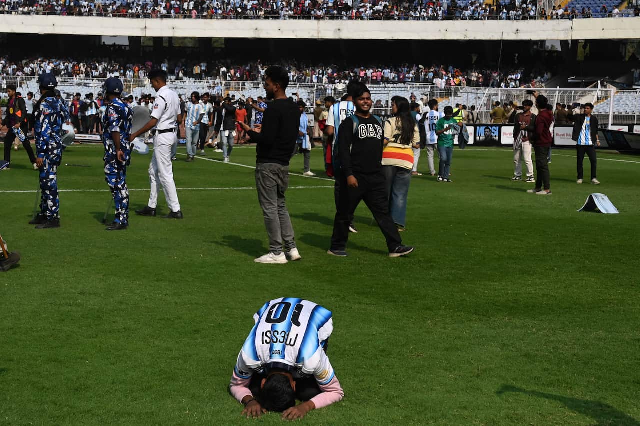 A man kneels down on a football field as a crowd gathers behind him